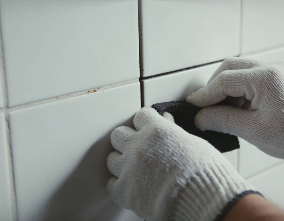 Mains gantees frottant joint sombre entre carreaux blancs.